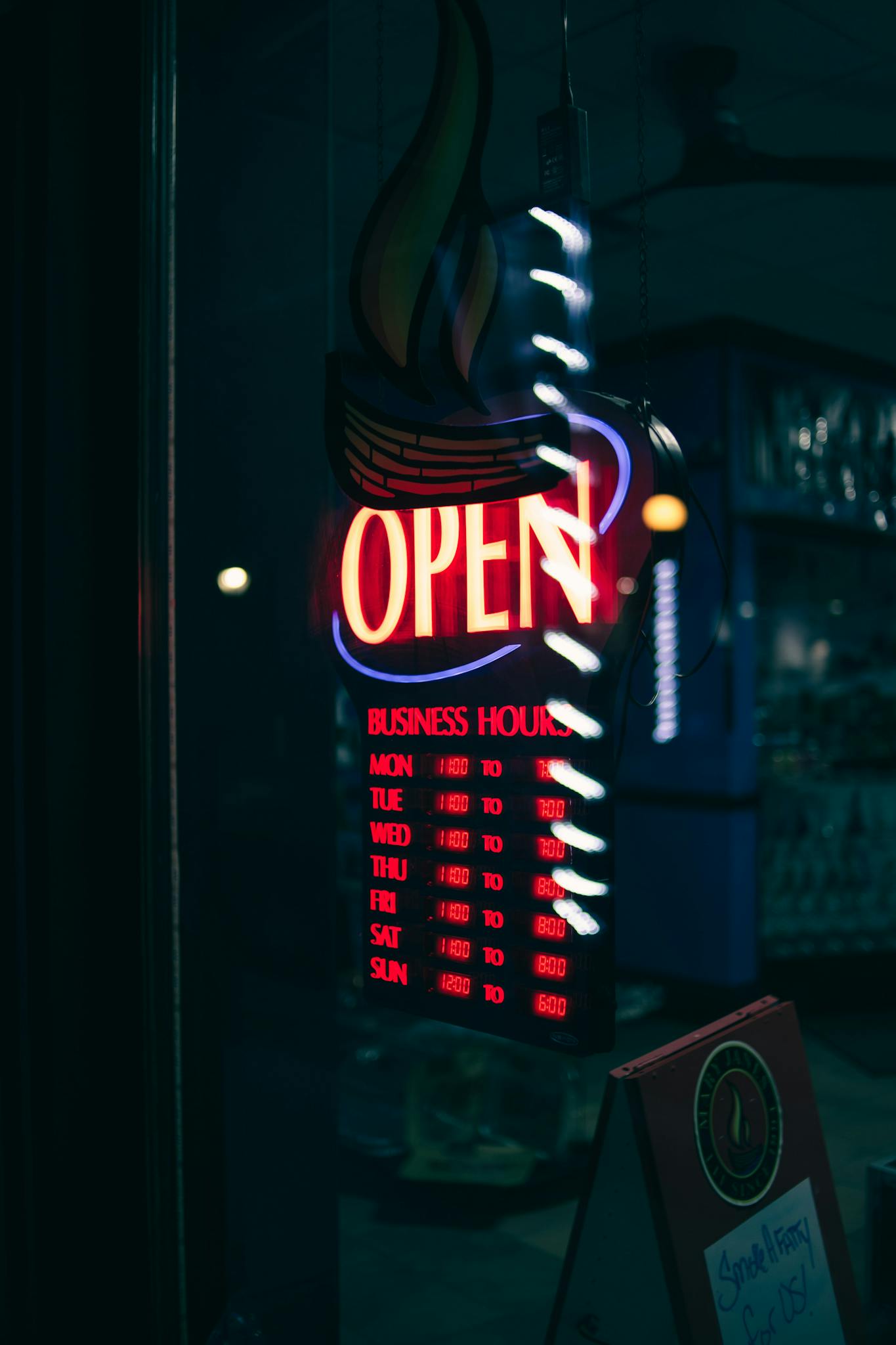 A vertical shot of a neon 'Open' sign displaying business hours in a dimly lit setting.