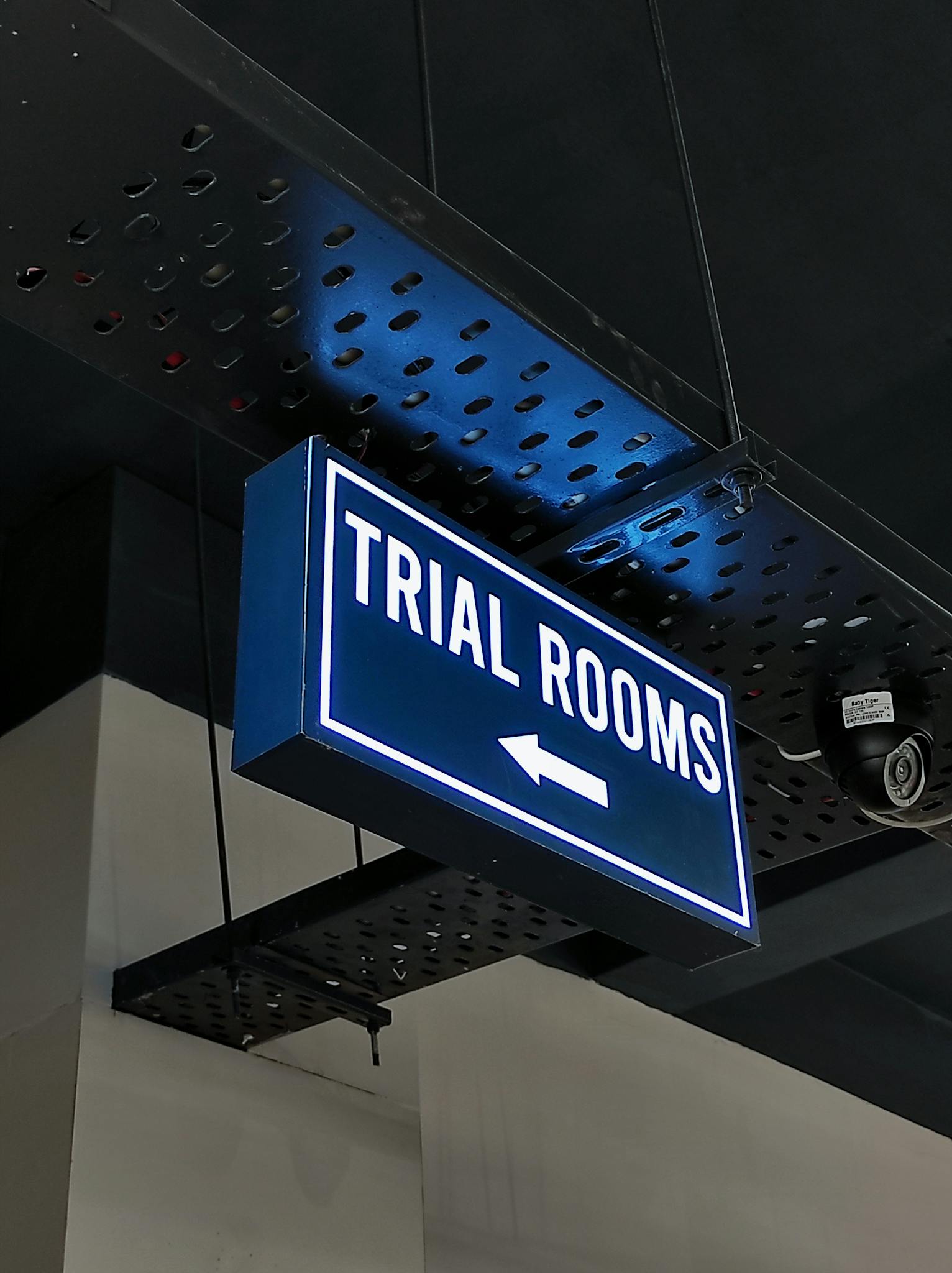 Blue and white trial rooms signage mounted on the ceiling indoors with CCTV nearby.
